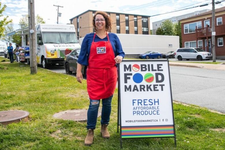 A woman with curly brown hair and glasses is posing and smiling for a photo. She is wearing a bright read apron with a blue blouse underneath. She poses by a sign that reads, 