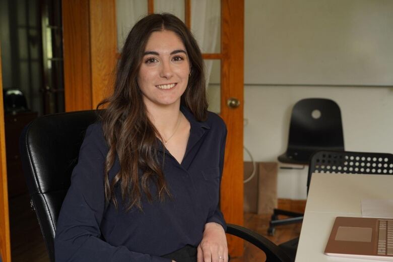 A woman in an office sits and smiles at the camera. 