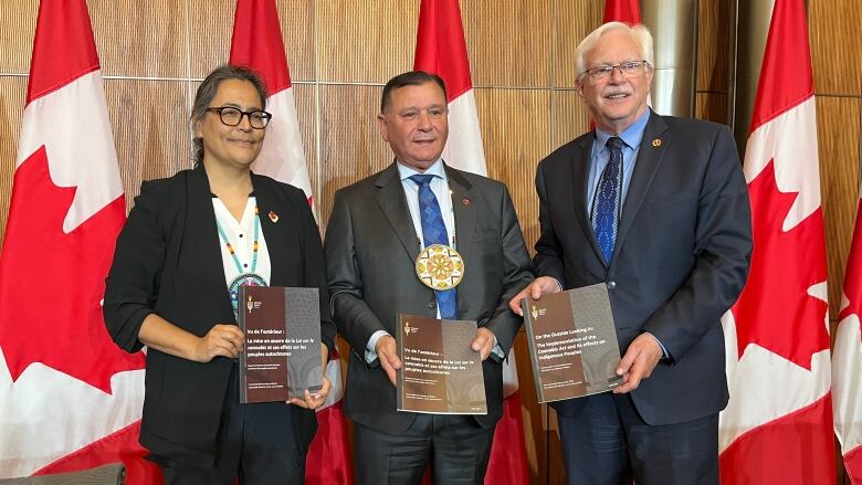 Three politicians holding a report in front of a row of Canadian flags.