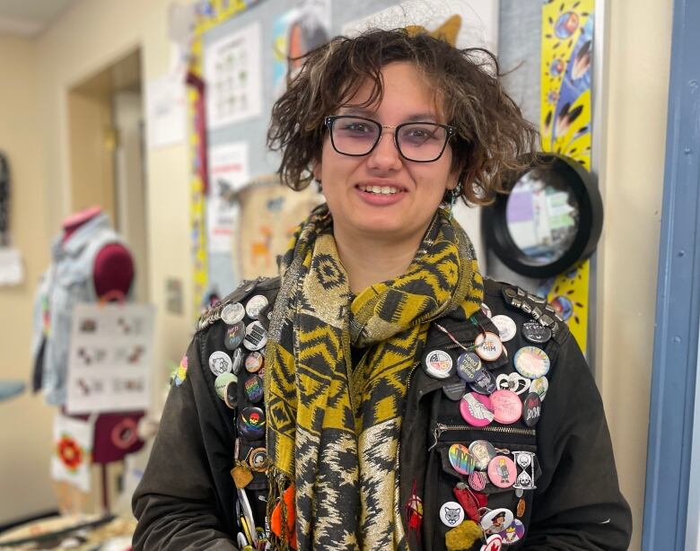 A grade 12 student with long short brown hair, large framed glasses wearing a yellow printed scarf and a jacket full of buttons. 