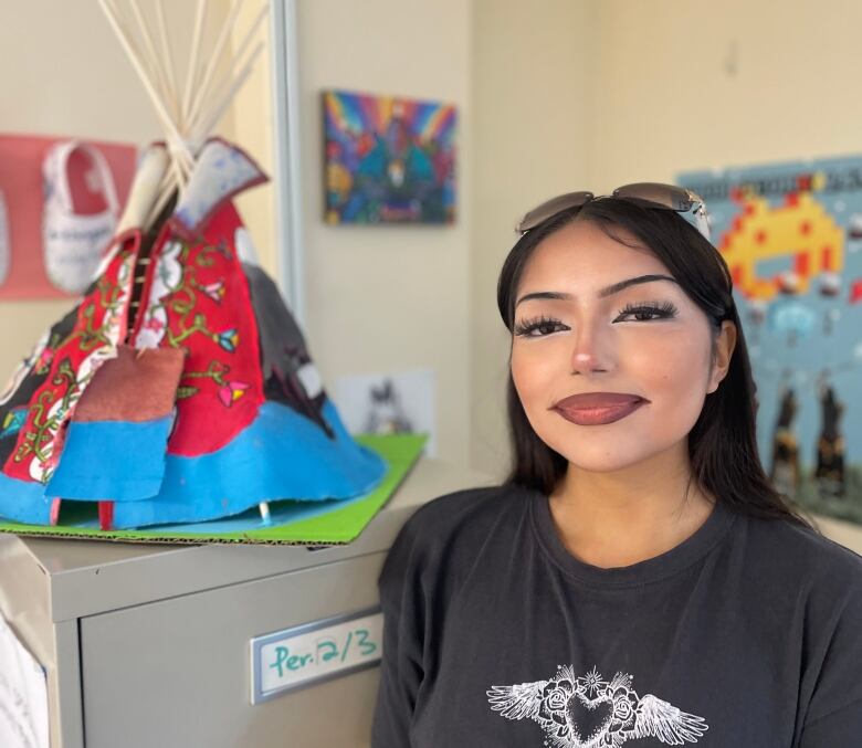 A grade 12 stuent with long black hair, red lipstick, and a black shirt is pictured standing next to a file cabinet with a tipi on top. 