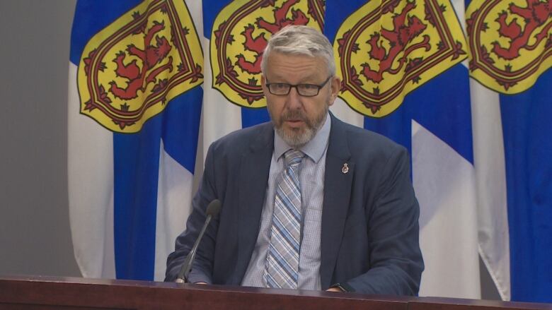 A man is seen sitting in front of the N.S. flag wearing black rectangular glasses, a navy blue suit jacket an a light blue gingham shirt.