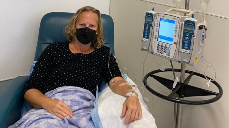 A blonde woman wearing a black mask sits in a chair while her left arm is linked to a treatment device. 