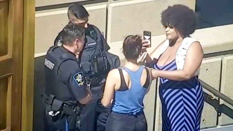 Two uniformed sheriffs stand with a woman in workout gear. Another woman with a megaphone stands near them and appears to be taking photos or video.