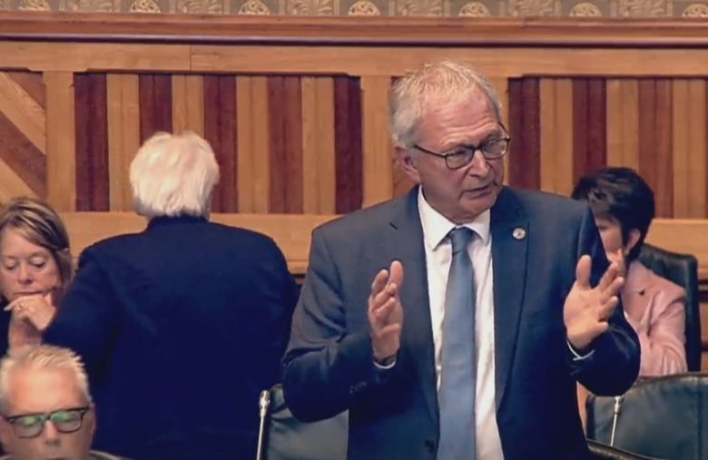 A man speaking at a podium surrounding by people sitting down. A woman on the left is standing with her back to the speaker.