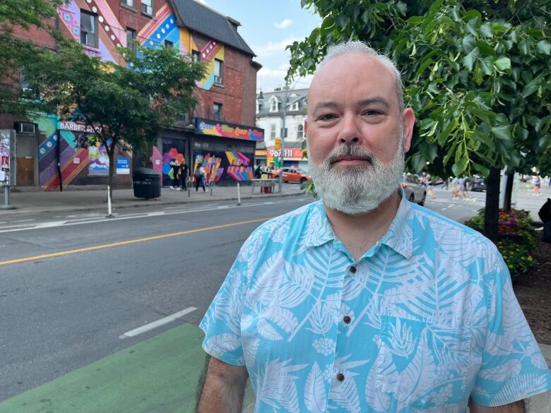 A tall man with silver hair and silver neatly trimmed beard stands on  a sidewalk wearing a blur floral print casual shirt.    