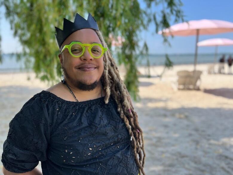 A Black man with long dreadlocks and bright green glasses stands near the lakeshore near a tree and with large pink sun umbrellas in the background. He's wearing a black shirt and a black leather crown. 