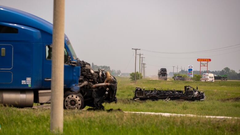 A semi trailer with a burned front end is pictured. A blackened passenger bus, destroyed by fire, is seen in the grass in the distance.