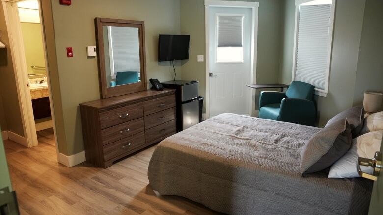 A room is shown to have a bed, dresser, tv, door, fridge, chair and small table, as well as bathroom door leading off to the side. 