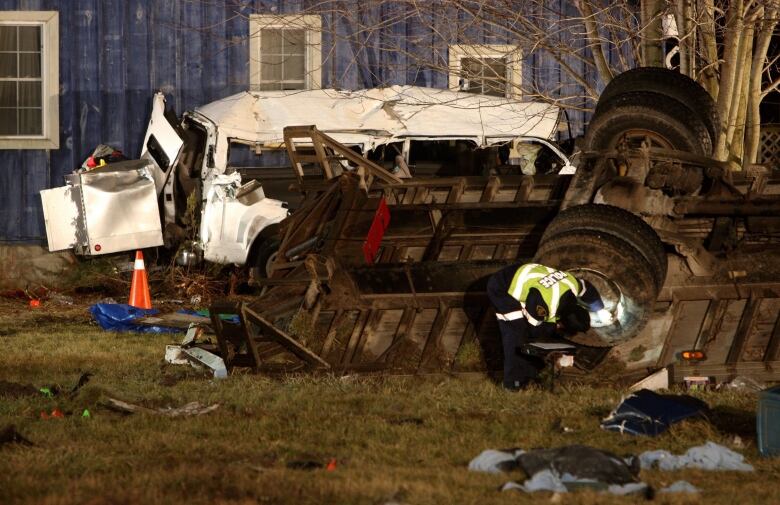 A crushed bus is seen on its side at night with a police officer bending over beside it.