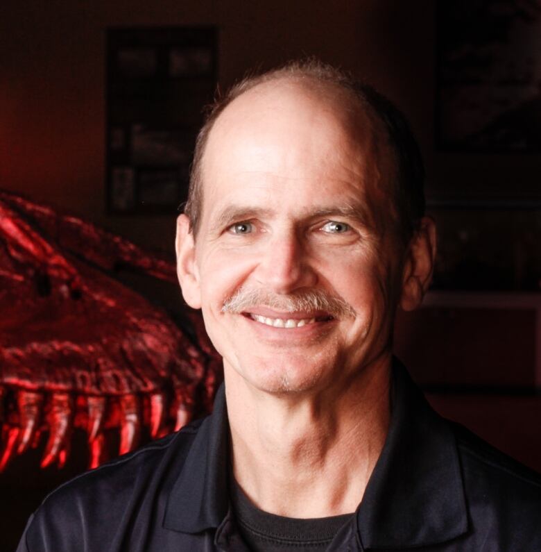 A man with a moustache smiles with the outline of dinosaur bones behind him. 