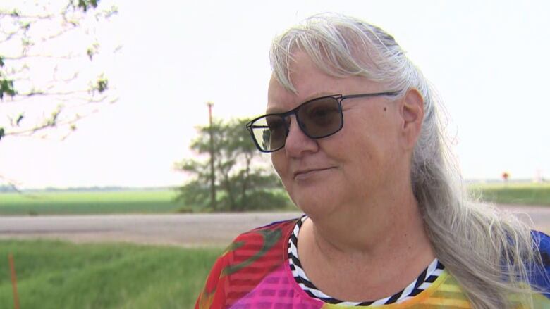 A white woman, with long white hair, is wearing sunglasses and a mutli-coloured shirt. She is standing near green fields, on the side of a dirt road.