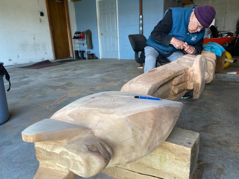 A older man sits in a garage carving a large totem pole.