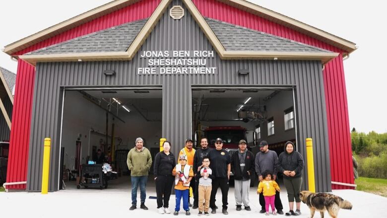 A red firehall with brown entrance is shown with a group of people standing in front of it. 