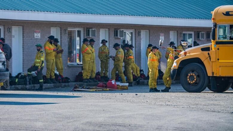 More than a dozen firefighters stand between a building and a yellow school bus. 