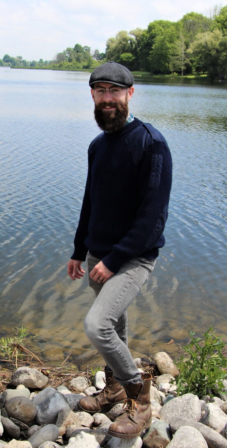 A man with a beard, eyeglasses and a grey page boy hat stands in front of a pond. He is wearing a blue sweater and smiling. 