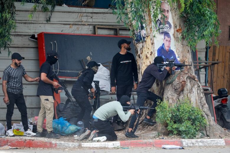 People wearing balaclavas aim their weapons while taking cover behind a tree.