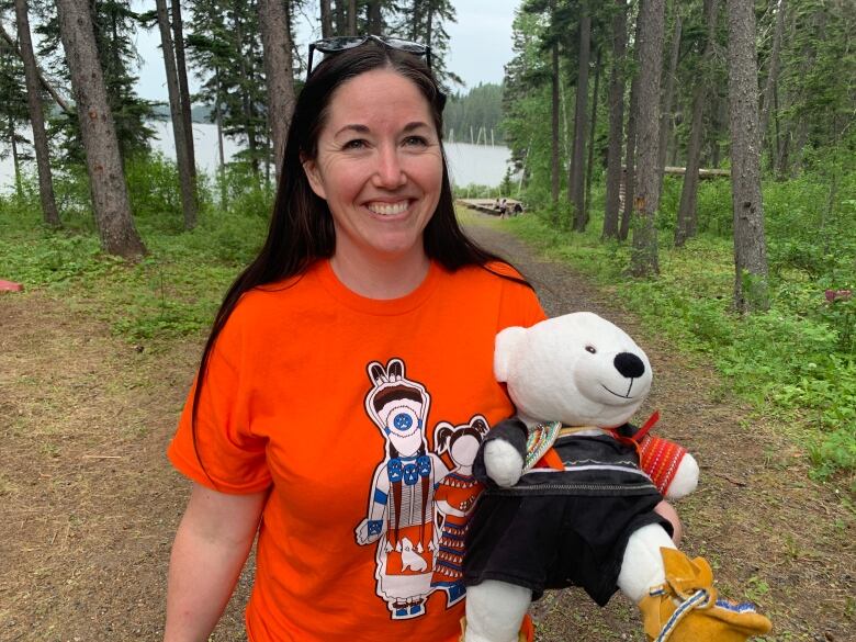 A woman with long dark hair in an orange shirt holds a white teddy bear wearing First Nations regalia.