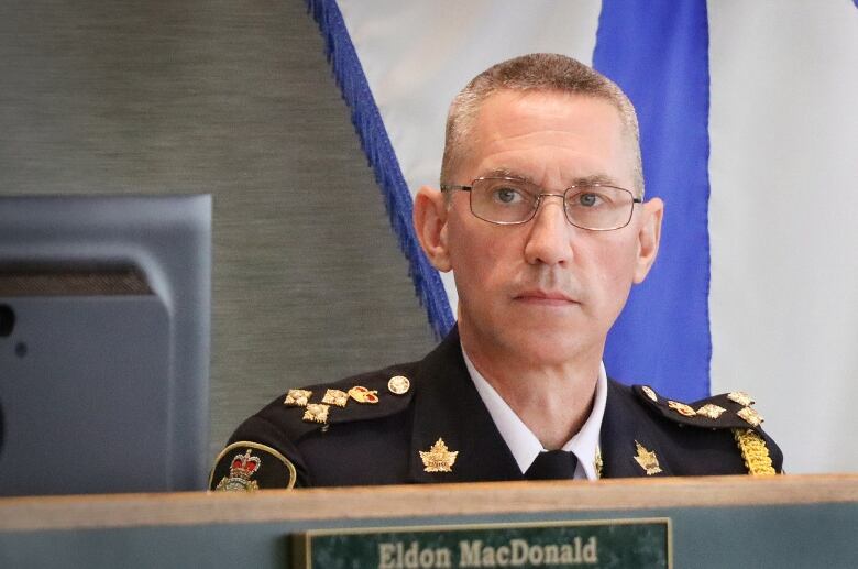 A man with short hair and glasses is in front of a Nova Scotia flag wearing a dark blue uniform with badges and pins on it.