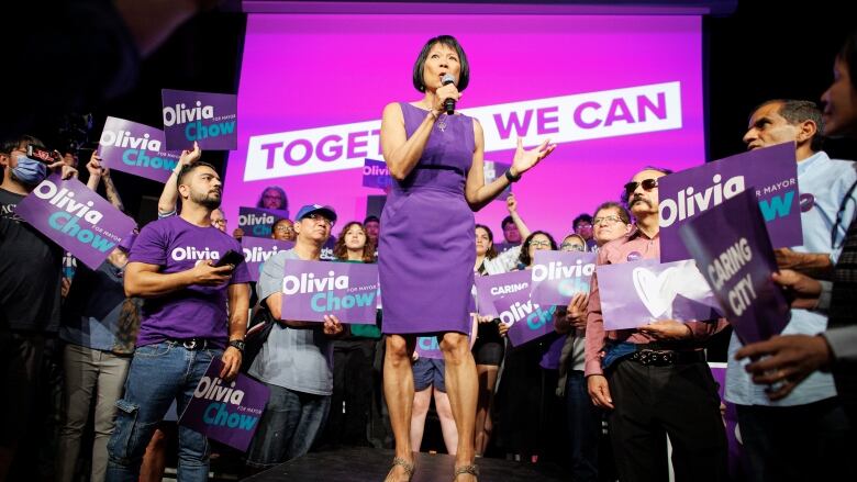 A woman on stage with supporters standing around holding signs. 