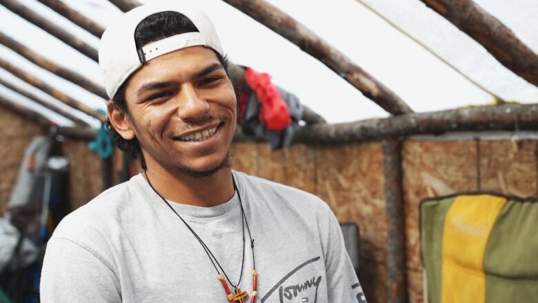 A man smiles at the camera. He's wearing a grey shirt and white hat. 