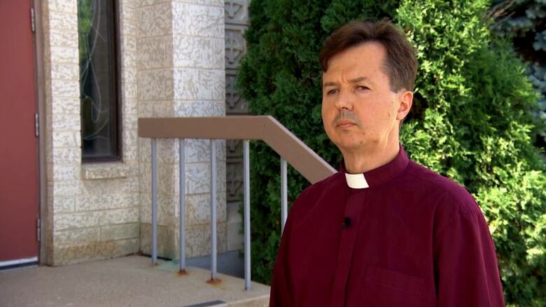 A man with a white priest's collar speaks to a reporter off-camera.