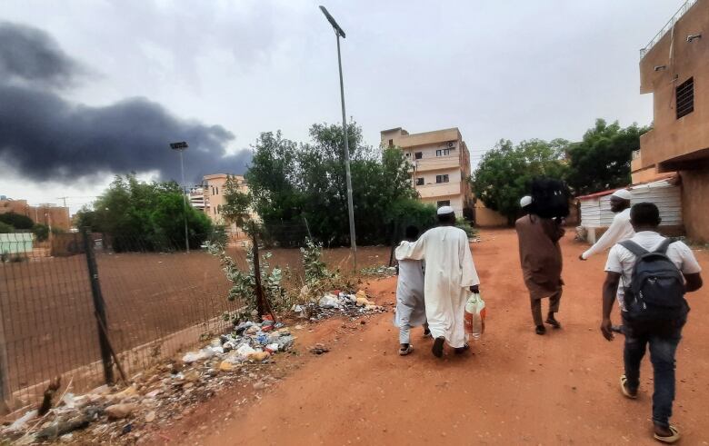 Black smoke rises above buildings, as people make their down a red dirt road in the foreground.