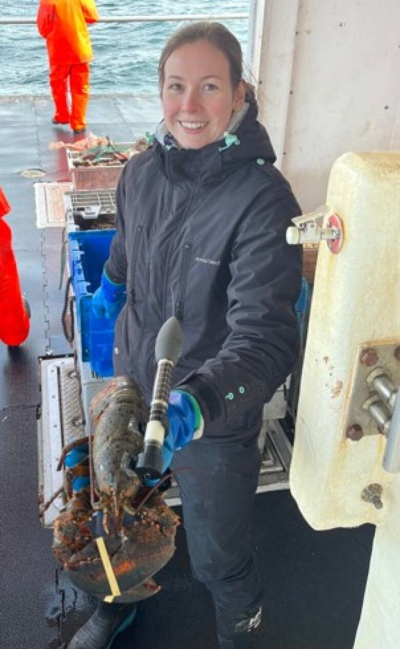 A woman wearing a coat on a boat holds a large lobster with a black tracker attached to it.