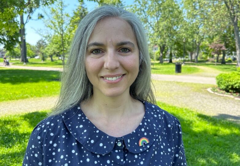 A woman with shoulder-length grey hair in a blue dress with a rainbow Pride pin smiling in a park with bright green trees and grass in the background.