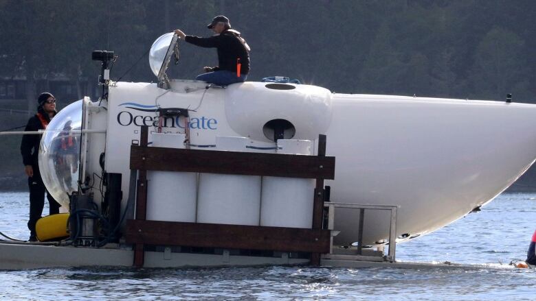 A man in a baseball hat emerges from the open hatch of a white submersible with the words OceanGate written on the side. 