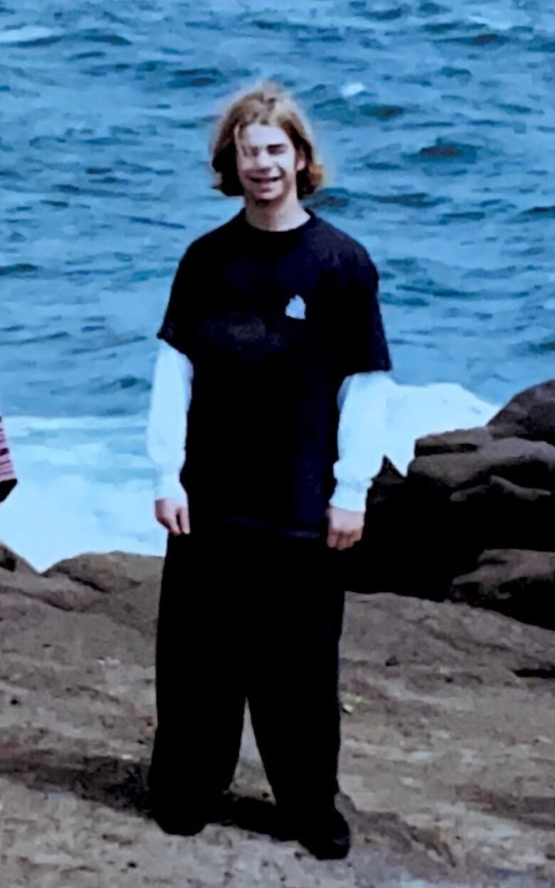 A young man with long hair and baggy pants smiles at the camera.