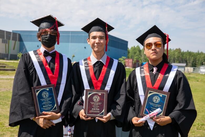 Three young men in graduation gowns hold plaques.