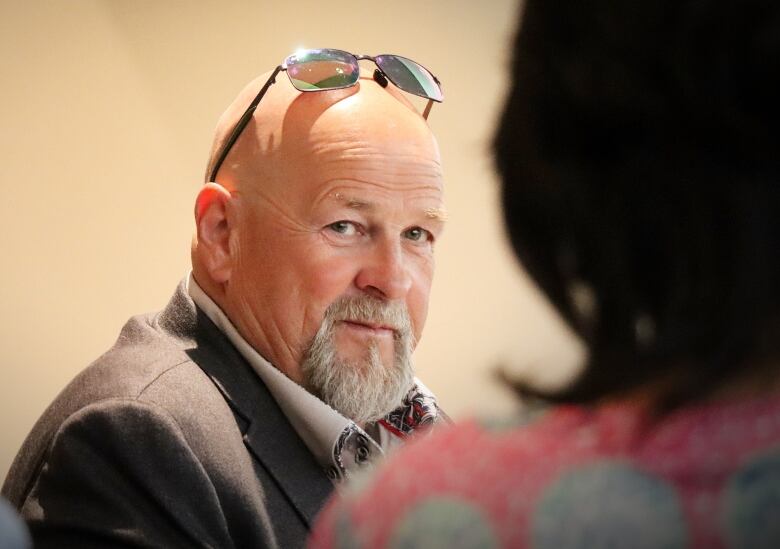 A bald man with a beard and moustache and sunglasses on his head speaks to someone.