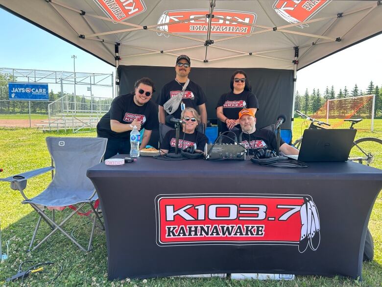 A group of people wearing matching merch shirts with the name of the K1037 station on them, smiling under an events tent. 