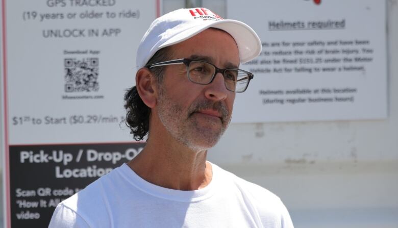 A white man with glasses and a beard wearing a white ball hat stands in front of a metal sea can