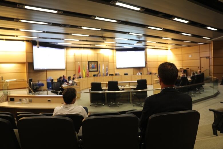 A man wearing a white shirt and another man wearing a black suit sit facing away from the camera, watching Regina city council. 