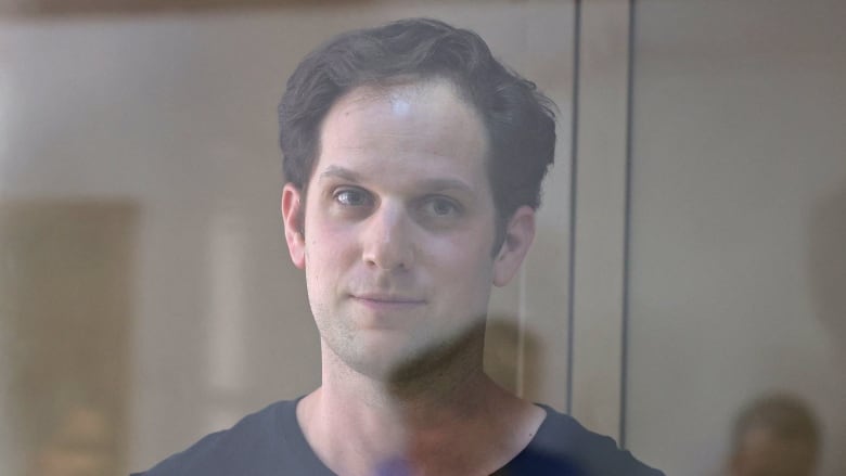 A clean-shaven man is shown in closeup through a glass partition.