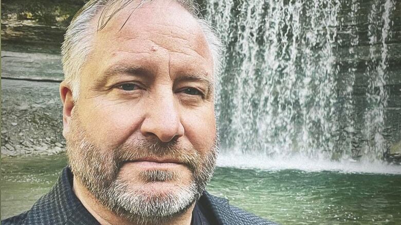 A man with a grey beard stands in front of a waterfall 