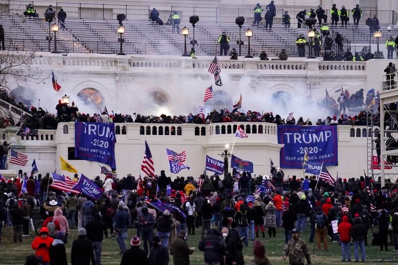 A wide shot is shown of hundreds of people gathered in a protest, with smoke rising in the air and flags held by some.