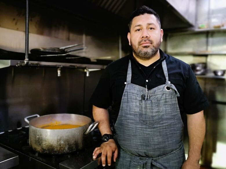 Chef Denis Hernandez stands in the kitchen wearing a black shirt and grey apron