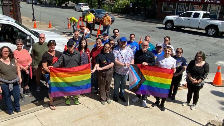 A group of about 20 people are smiling, with two large Pride flags held up front. There are workers in the background working on the Pride crosswalk.