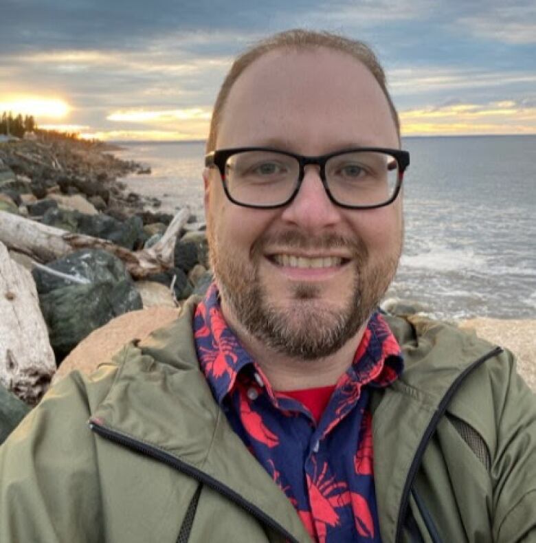 A man with a beard and glasses is smiling. The ocean, rocks, and logs are in the background. The man is wearing a windbreaker and collared shirt.