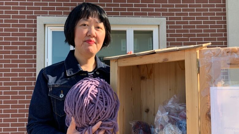 A woman holds a ball of yarn beside a wooden box filled with more yarn.