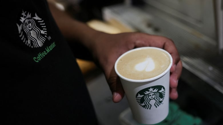 The hand of a Starbucks barista preparing a drink with a heart in the foam is shown.