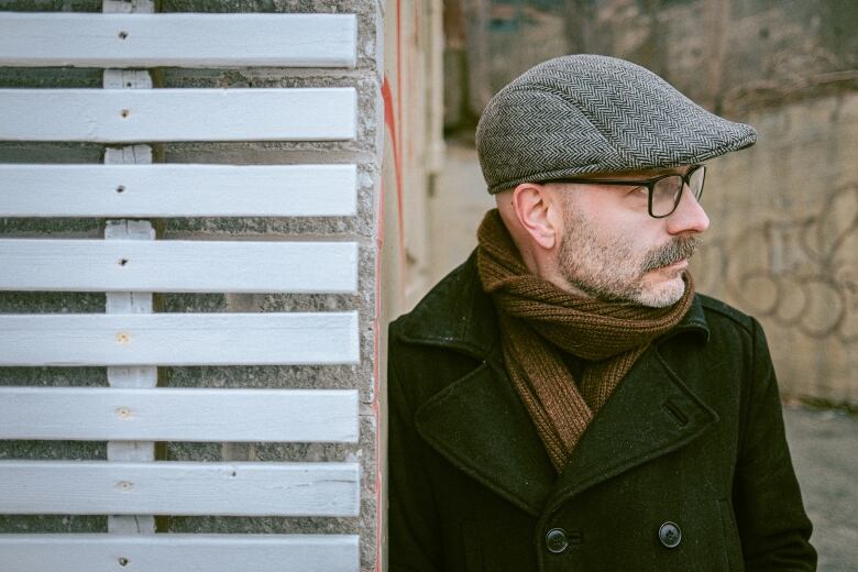 A man wearing glasses and a hat leaning against a wall. 