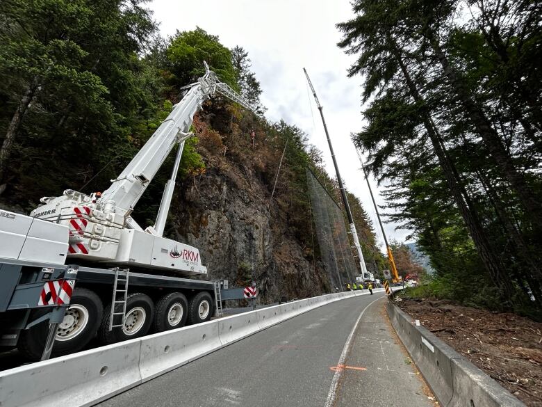 A crane truck helps install protective netting on a rock face beside a closed highway as workers operate on the roadway.