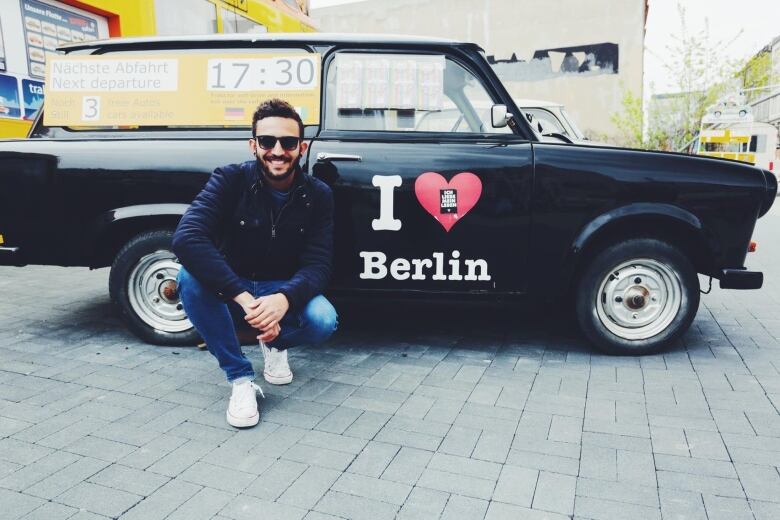 A person with short hair and sunglasses crouches in front of a Berlin taxi.