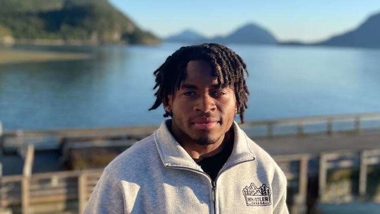 A man stands in front of body of water with mountains in the background.