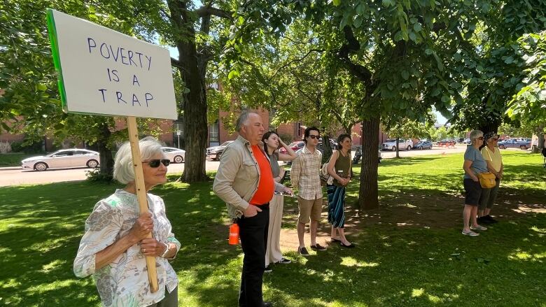 A small group of people gather under trees outside the provincial legislature. One holds a sign that says 'poverty is a trap.'