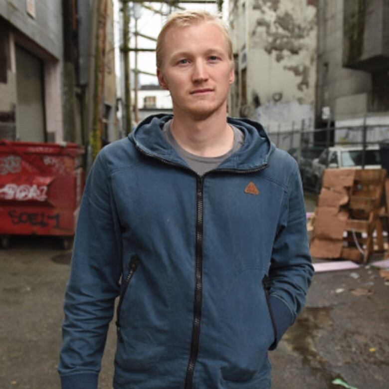 A man in a blue hoodie stands in an urban alley way.